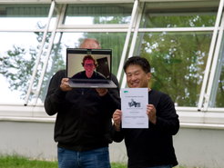 Zwei Personen mit einem Laptop und einem Preis-Zertifikat vor einem Gebäude.